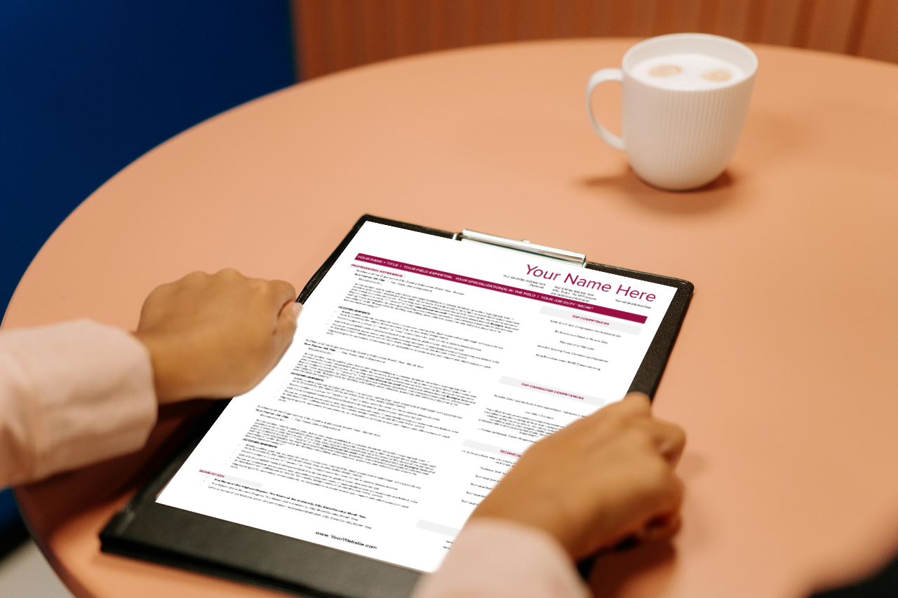 A resume on a clipboard gripped by two hand of a person sitting at a coffee table with a white mug in front of them.