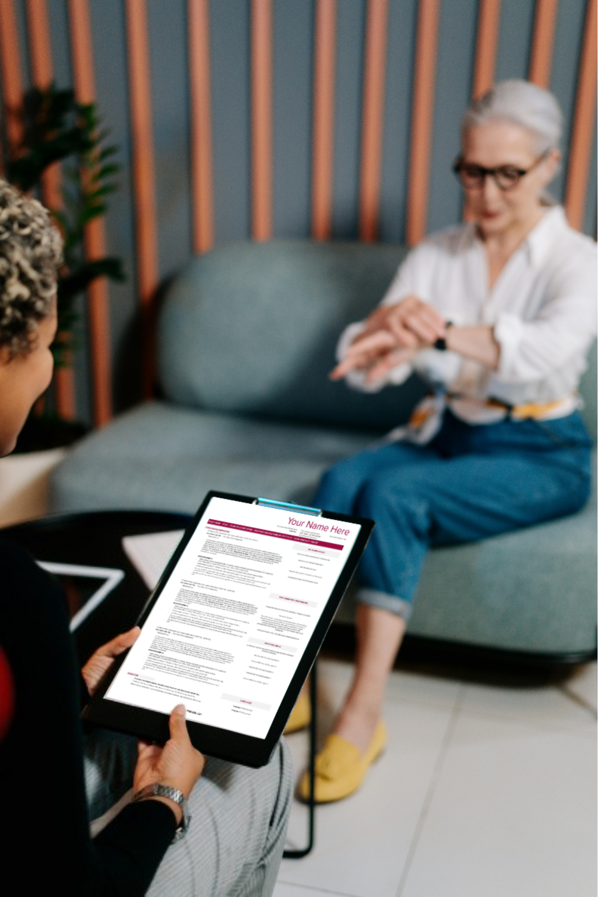 A woman holding a clipboard with a resume to interview another woman sitting across from her on a sofa.